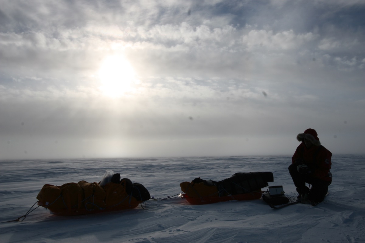 Polar traveller kneeling beside two loaded sledges on an ice sheet under a low sun
